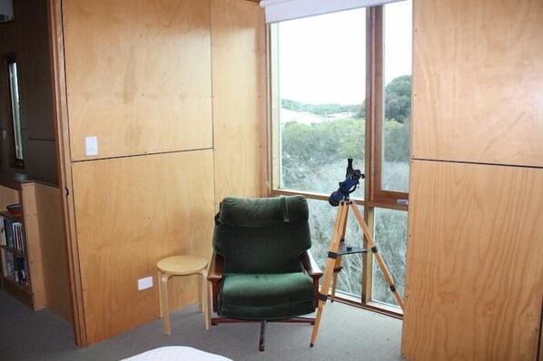 Una habitación con una silla verde, un taburete pequeño y un telescopio cerca de una ventana que ofrece vistas a árboles y colinas.