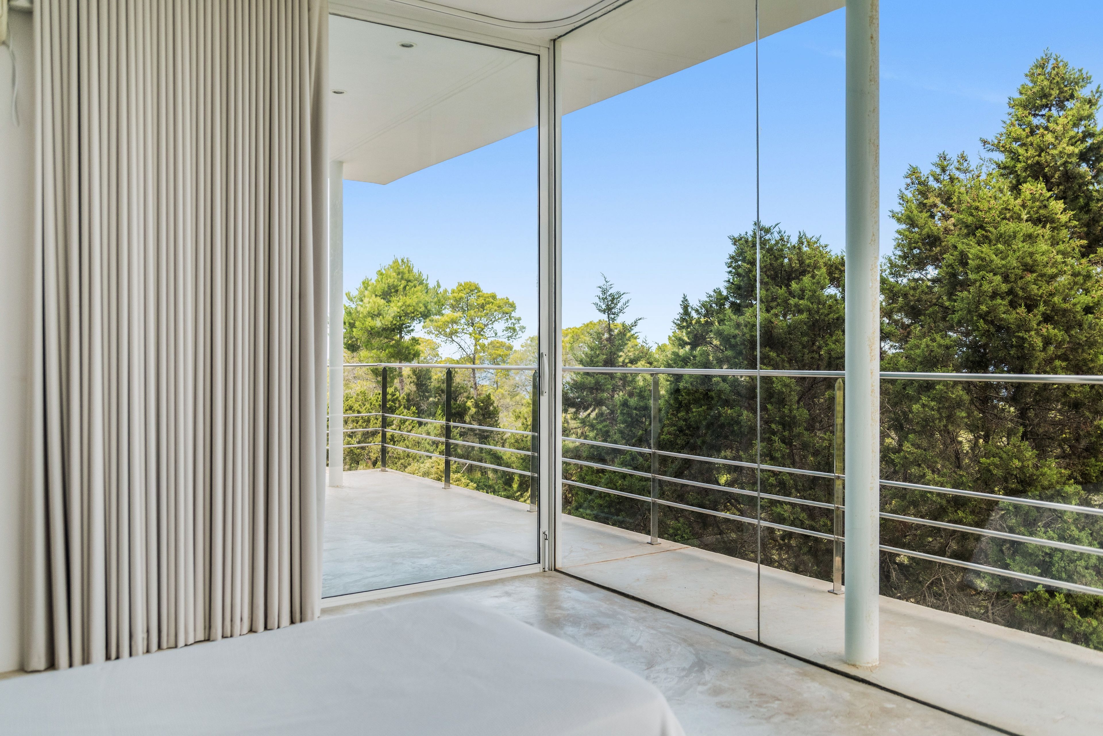 Una habitación moderna con un amplio balcón de vidrio, barandilla metálica y vistas a árboles.
