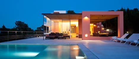 A modern house with a swimming pool, outdoor seating, and a well-lit interior.