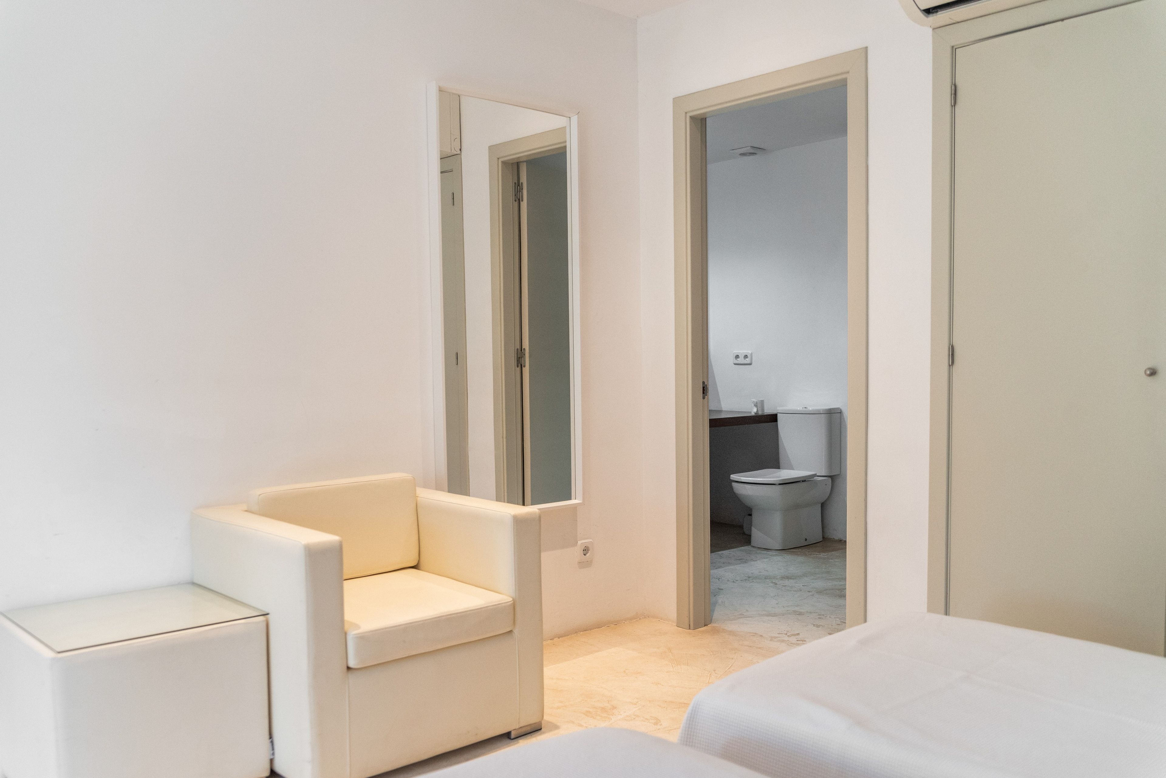 Een moderne slaapkamer met een witte stoel, een bed en een badkamer zichtbaar door een open deur.