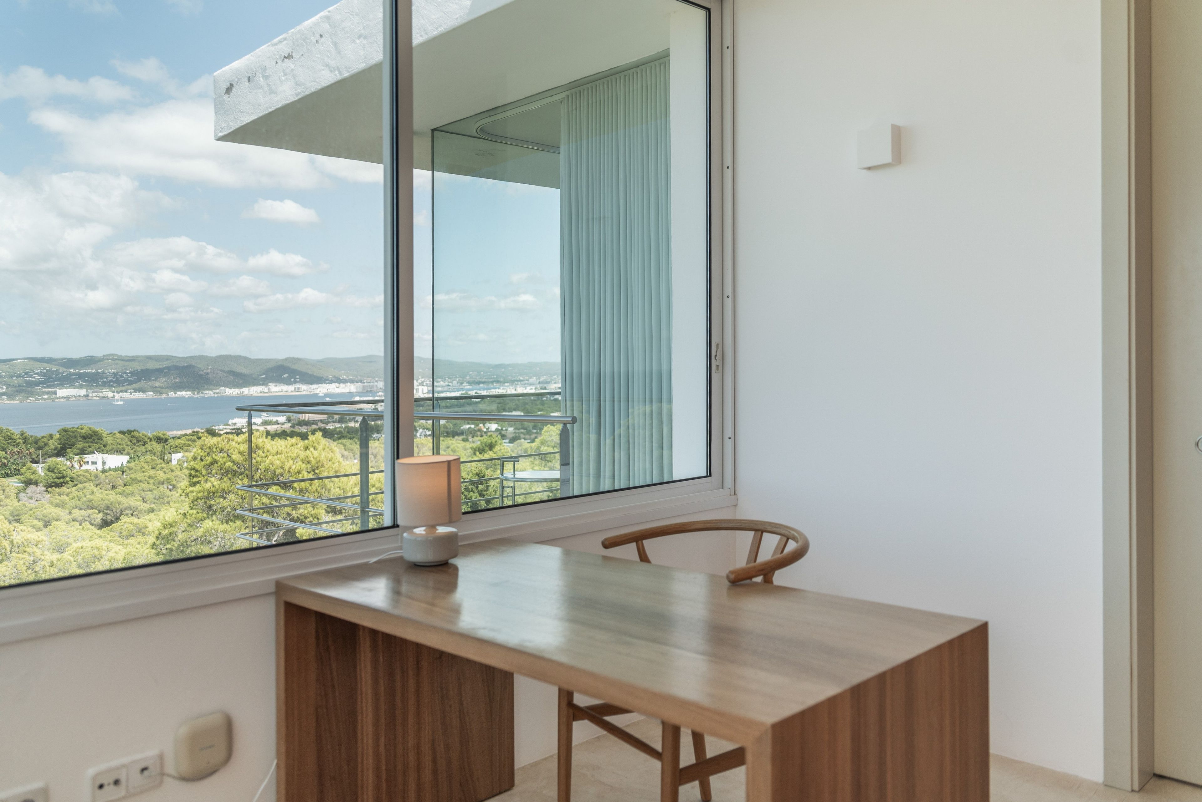 A room with a view of a body of water and hills, a wooden desk with a chair, and a window with blinds.
