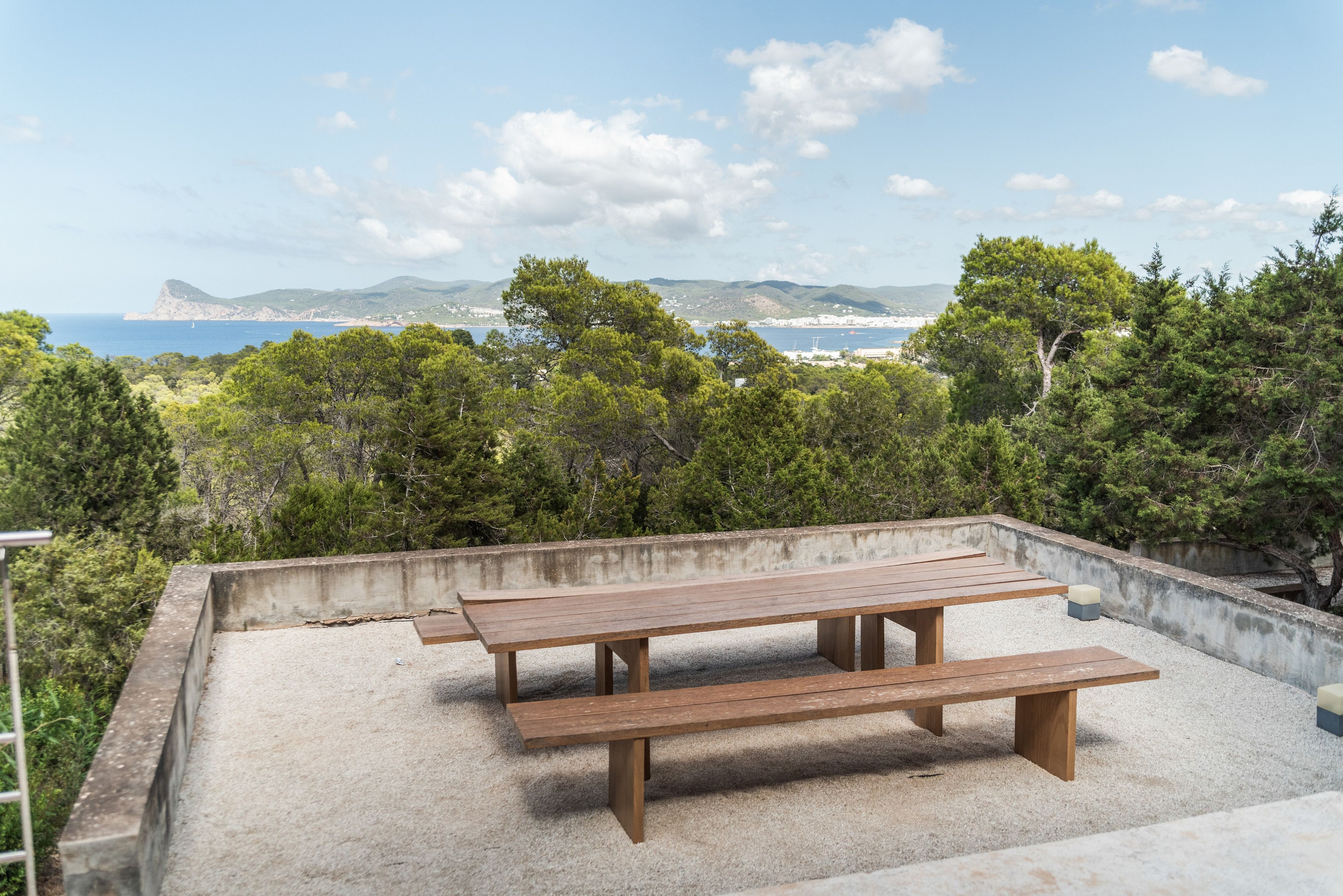 Une terrasse en béton avec deux bancs en bois, donnant sur un paysage côtier composé d’arbres et de collines.