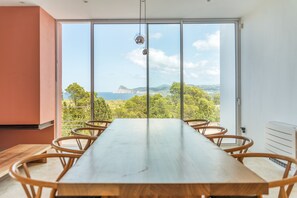 A modern dining area with a long table and chairs, large windows offering a view of a coastal landscape.
