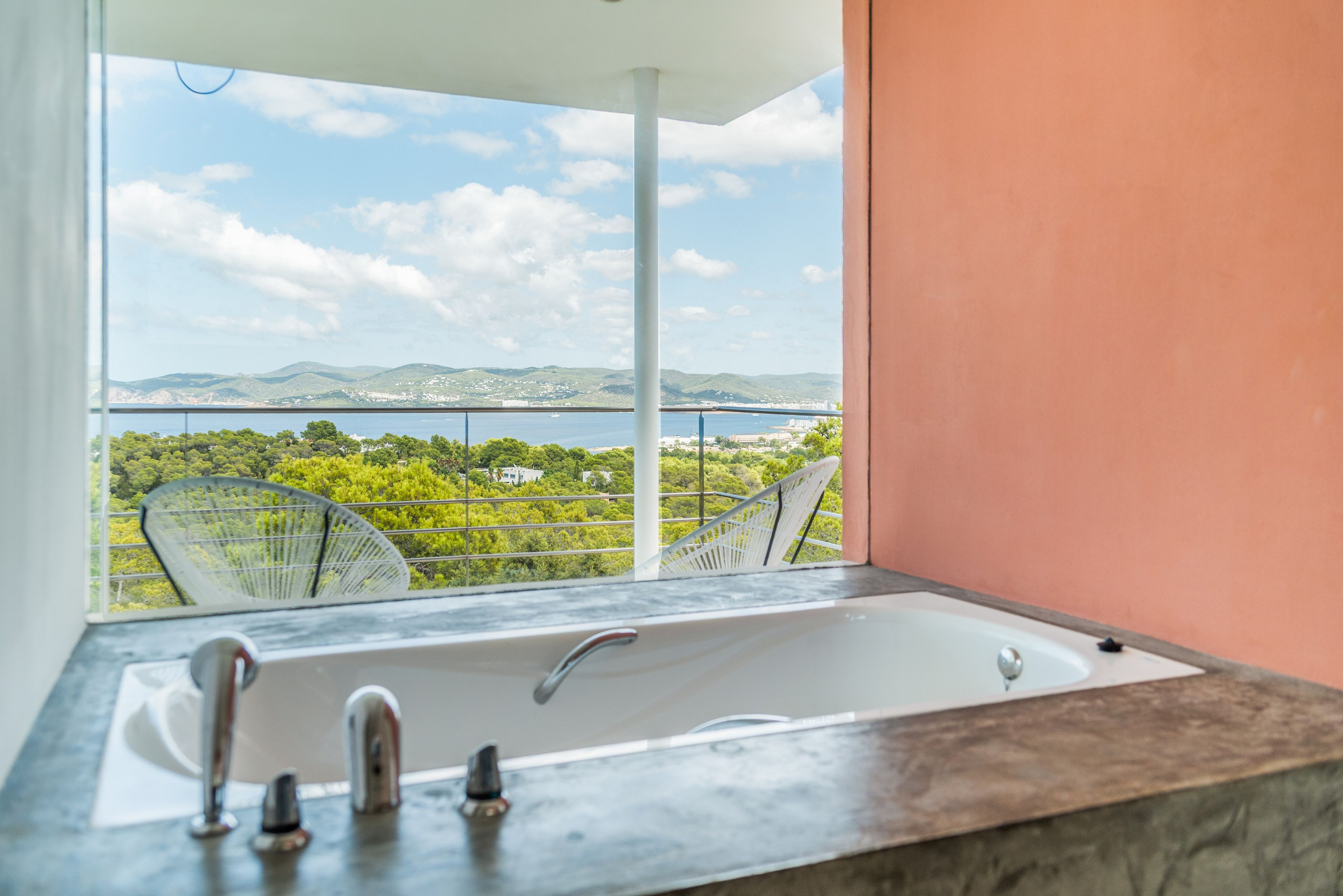 Baño con bañera, dos grifos y vista a un paisaje costero con árboles y colinas.