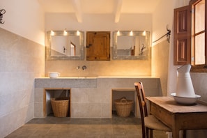 A bathroom with a wooden door, two mirrors, a sink, a chair, and a table with a white pitcher.