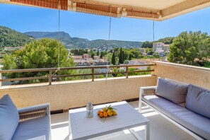 A balcony with a white sofa, a small table with a fruit bowl and a lantern, and a view of a town and mountains. - Fantastic Apartment "Puerto Rico Port de Sóller" with Mountain View, Terrace and Wi-Fi (Port de Sóller)