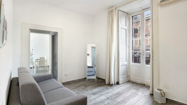 Ein modernes Wohnzimmer mit einem grauen Sofa, einem Spiegel und Blick durch eine Tür auf einen Außenbalkon.