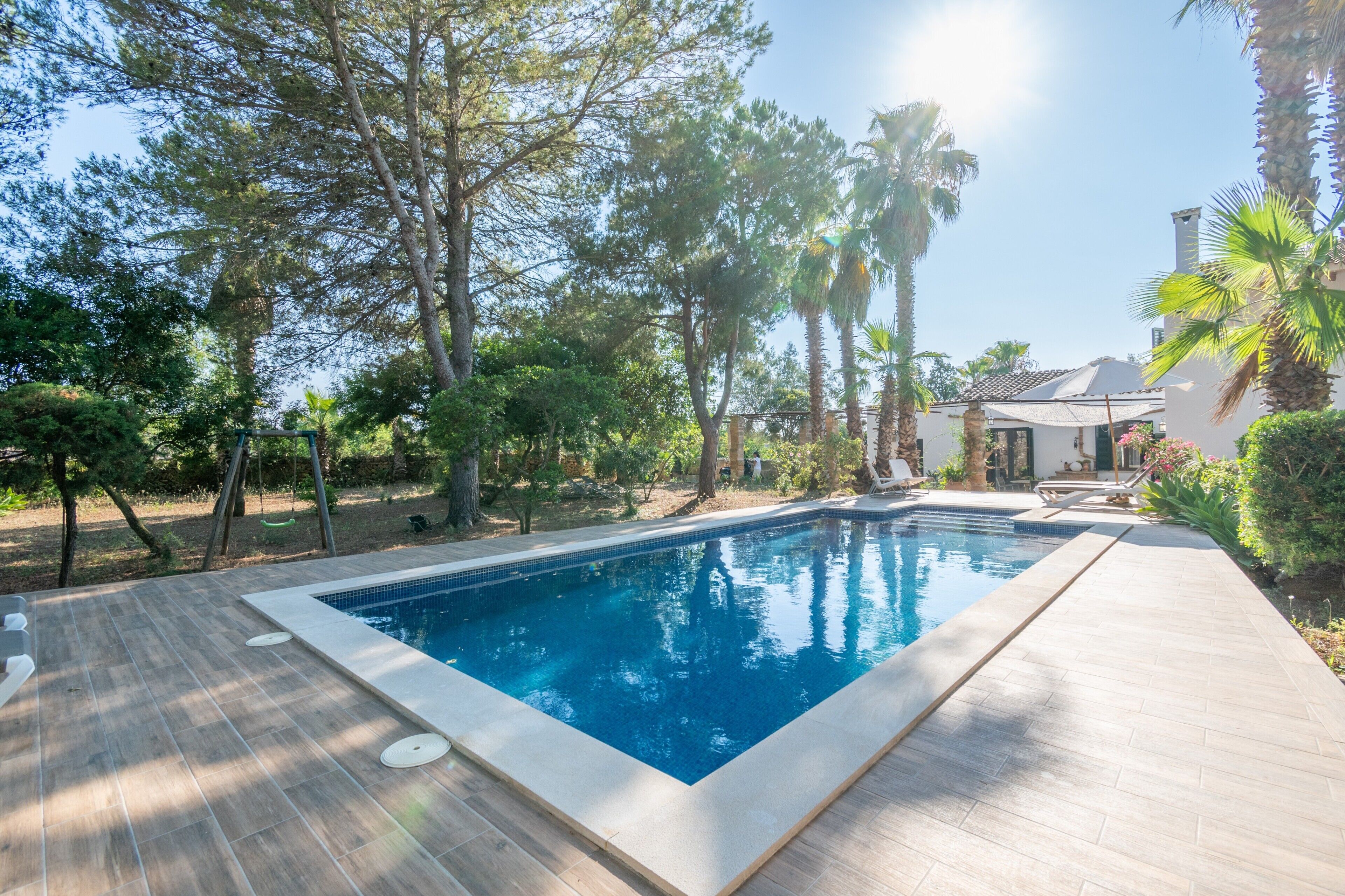 A rectangular swimming pool with a tiled deck, surrounded by trees and a house in the background.