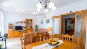 Ein Essbereich mit Holztisch, Stühlen und einer Obstschale. Ein Fernseher steht auf einer Holzkommode, neben einem Sofa und einer Lampe.