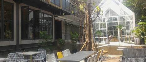 Un área de comedor al aire libre con mesas y sillas, rodeada por un edificio con ventanales grandes y una estructura de vidrio al fondo.