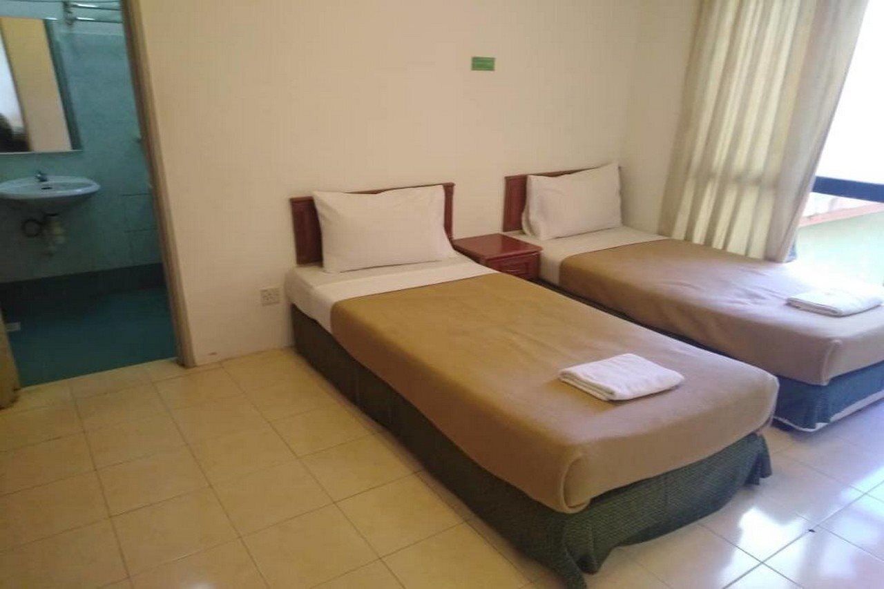 Habitación de hotel con dos camas individuales, una mesita de noche pequeña y un baño con lavamanos y ventana con cortinas.