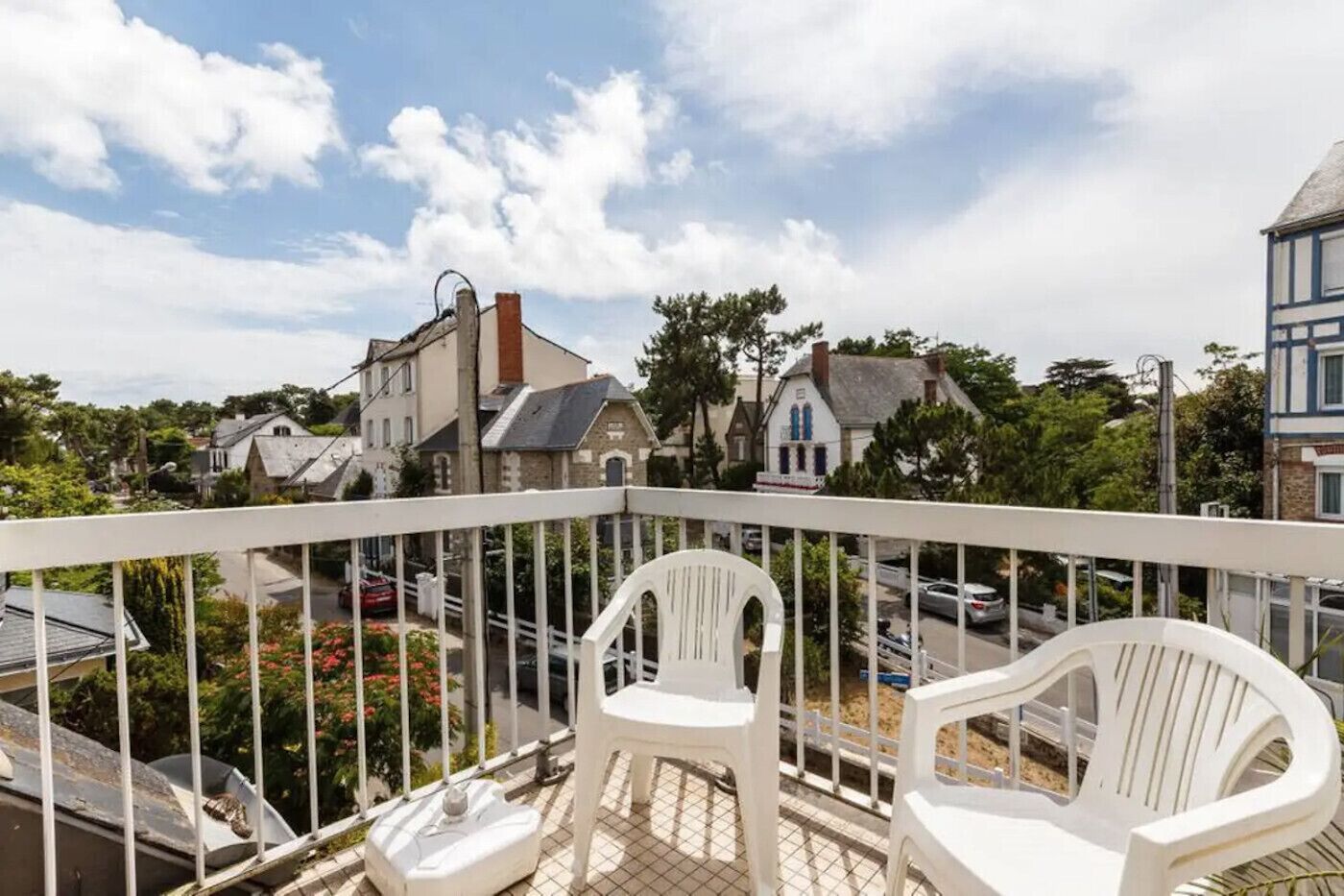 A balcony with white chairs, a view of a street with houses, and a clear sky.