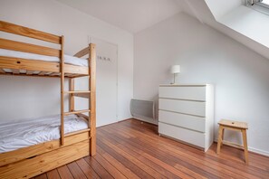 A bunk bed with a wooden frame and white bedding, a white dresser with drawers, a small wooden stool, and a radiator on the floor.