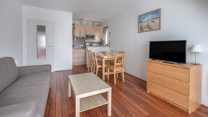 Een knus woongedeelte met een bank, een eettafel, stoelen, een televisie op een houten kast, en een keuken met kastjes en een magnetron.