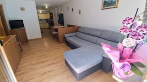 A modern living room with a grey sectional sofa, wooden flooring, a dining area with a table and chairs, and a kitchen in the background.
