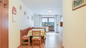 Un comedor con banco y mesa de madera, una cocina al fondo y vistas a través de una ventana.
