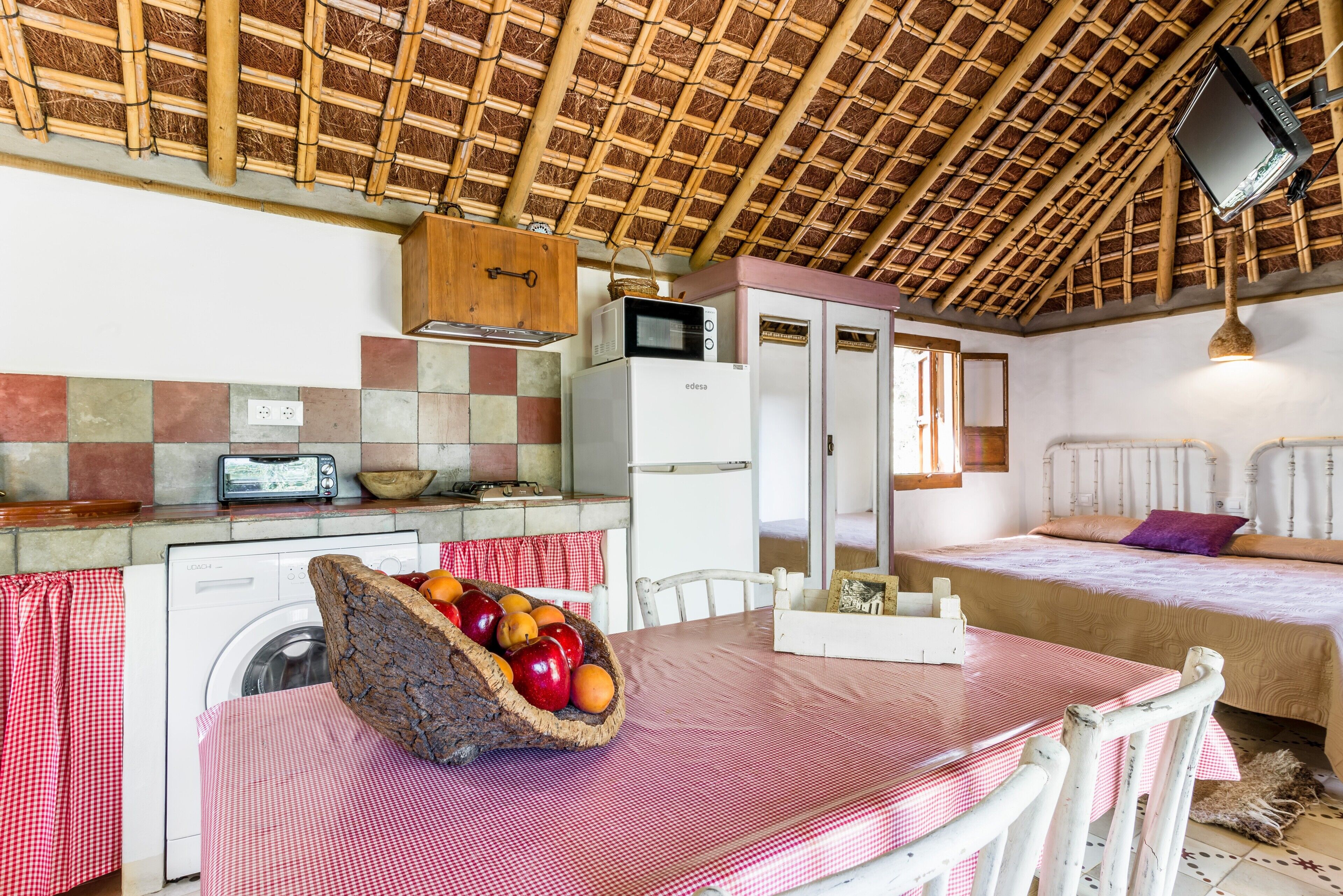 A compact kitchen with a washing machine, a microwave, and a refrigerator. There is a dining table with a basket of fruit and a red and white checkered tablecloth.