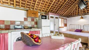 A compact kitchen with a washing machine, a microwave, and a refrigerator. There is a dining table with a basket of fruit and a red and white checkered tablecloth.