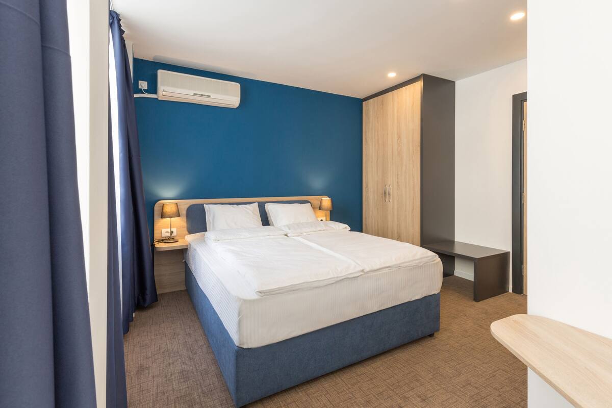 Una habitación de hotel moderna con una cama grande, una mesita de noche con lámpara, un armario y un equipo de aire acondicionado en la pared.