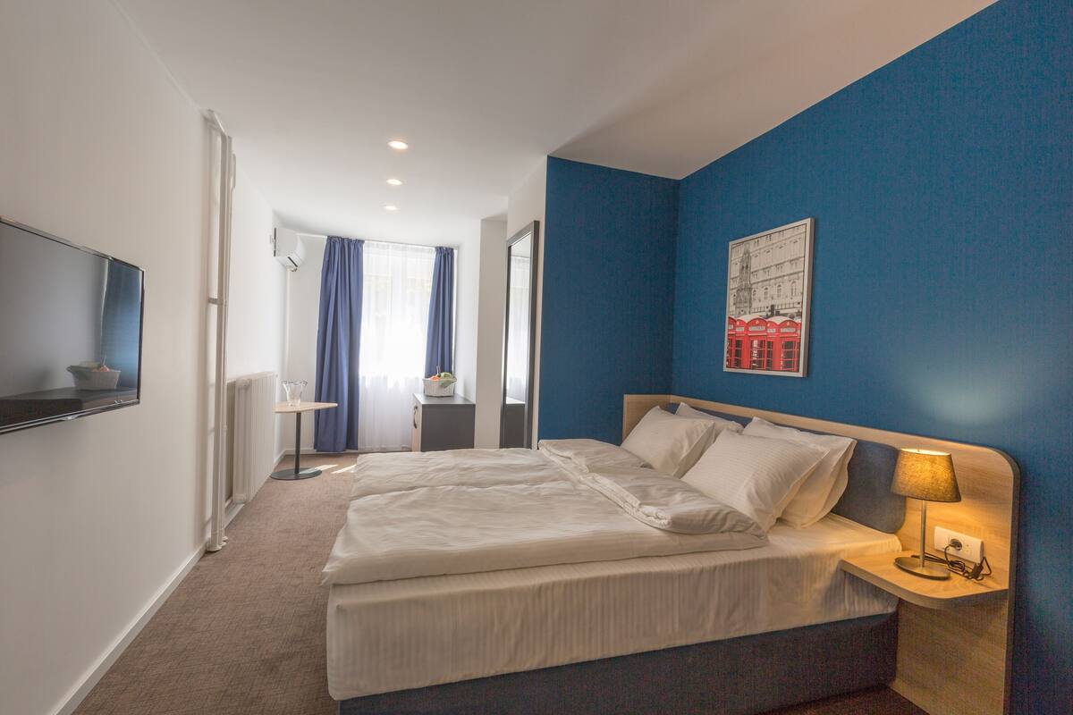 Habitación de hotel con una cama grande, una mesita de noche con lámpara, un televisor de pantalla plana en la pared y una ventana con cortinas.