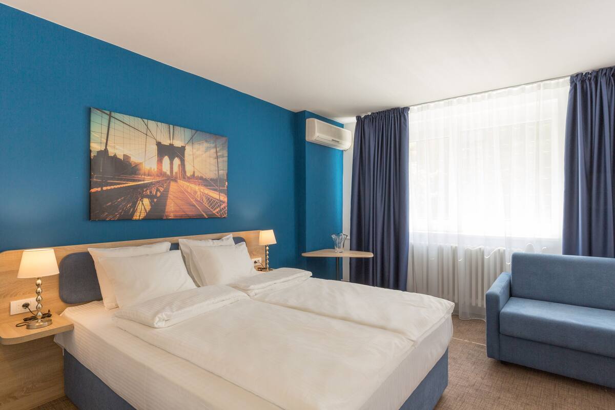 Una habitación de hotel con una cama grande, un sillón azul, una mesita y un cuadro en la pared.