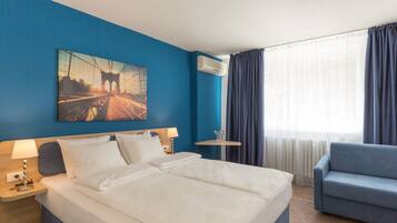 A hotel room with a large bed, a blue armchair, a small table, and a painting on the wall.