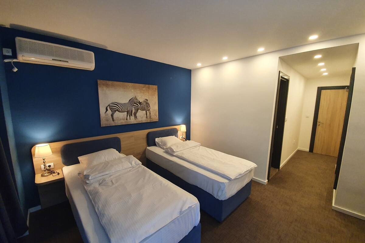 Habitación de hotel con dos camas, aire acondicionado instalado en la pared, un cuadro de cebra y una puerta que da a otra habitación.
