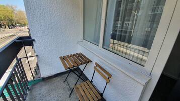 A balcony with two wooden chairs and a small table, a white textured wall, and a large window overlooking a building.