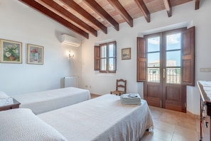 A room with two beds, wooden beams on the ceiling, a window with wooden shutters, and a door leading outside.