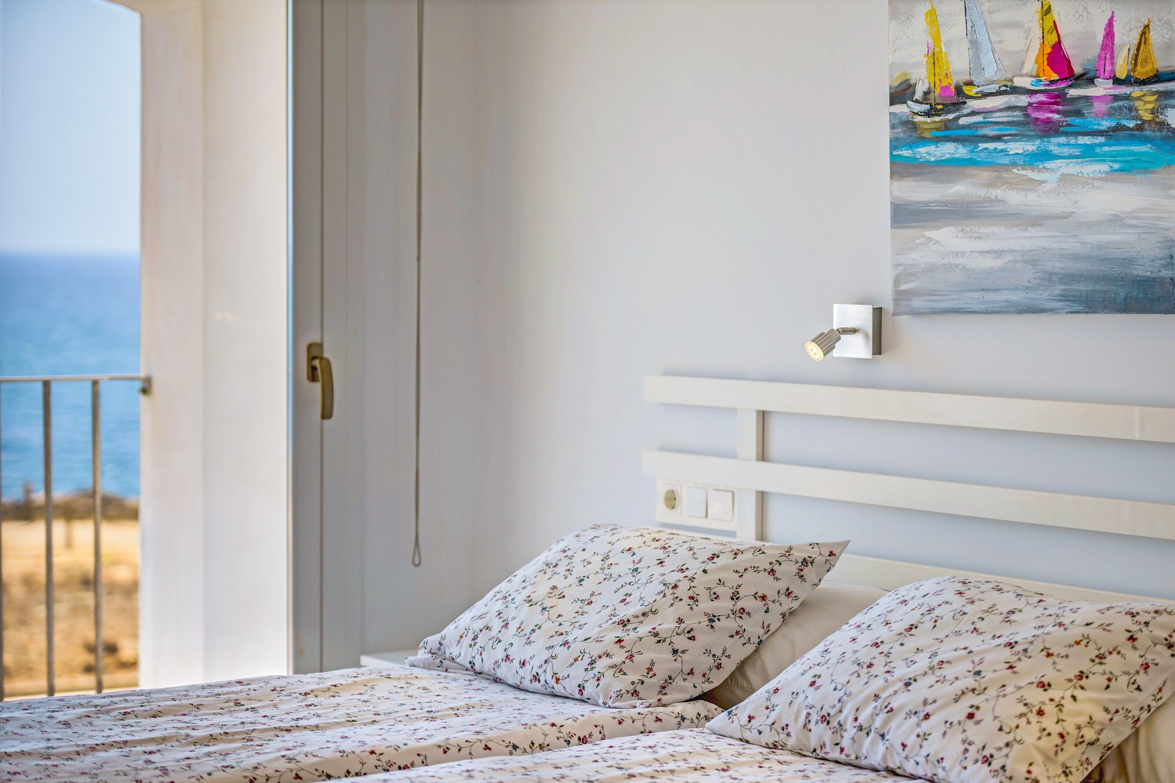 Une chambre à coucher avec un lit, des oreillers, un tableau représentant des voiliers et une vue sur la mer.
