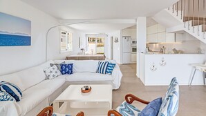 Een moderne woonkamer met een bank, salontafel en houten stoelen. In de achtergrond is een keuken te zien met een wit aanrecht en kastjes.