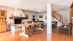 Un vaste espace de vie comprenant une grande table en bois, plusieurs chaises, une cheminée, un escalier et un coin salon confortable.