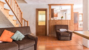 Un salon avec un escalier en bois, un canapé avec des coussins, un fauteuil à motifs et un coin cuisine visible à travers une porte ouverte.