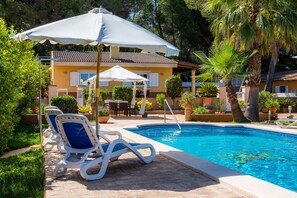 Zona de piscina con sillones de descanso, sombrillas y un edificio con techo de tejas.