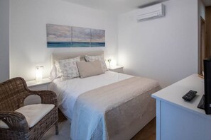 A bedroom with a bed, a nightstand, a wicker chair, a desk with a remote control, and a wall-mounted air conditioner. - Duplex Flamingo Cadiz (Cádiz)