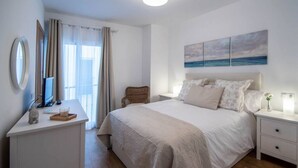 A bedroom with a large bed, a bedside table with a lamp, a chair, a desk with a computer, and a window with curtains. - Duplex Flamingo Cadiz (Cádiz)