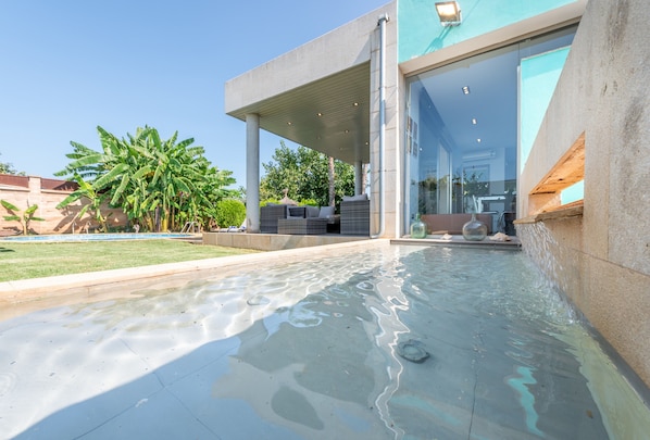 Un área de piscina moderna al aire libre con una zona de estar techada, un espacio con paredes de cristal y un cielo despejado.
