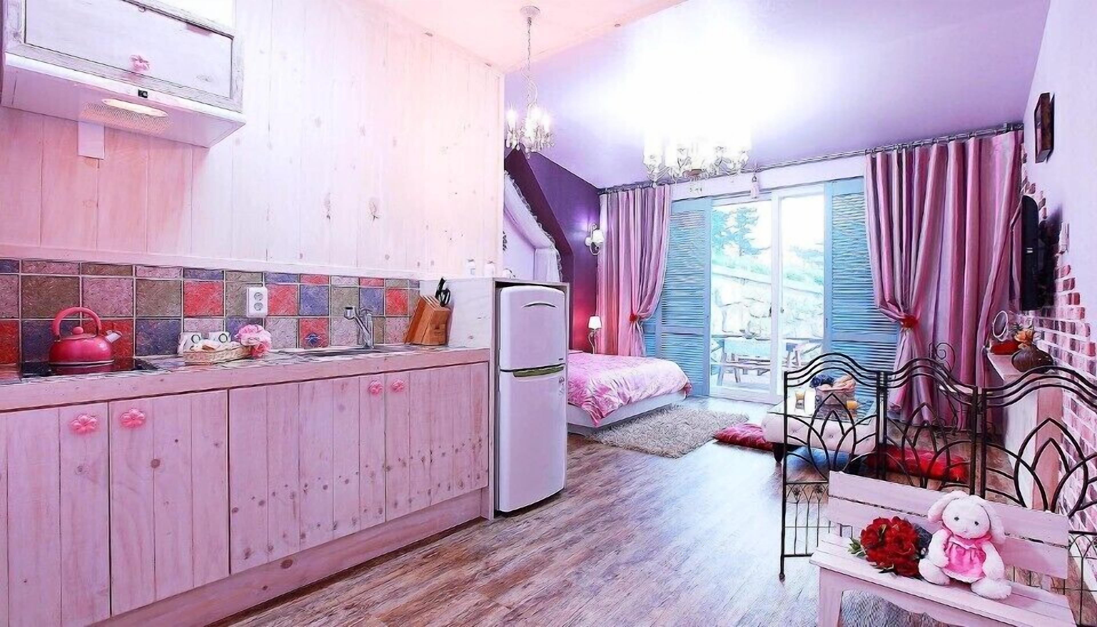 Ein zartrosa eingerichtetes Zimmer mit Küche, einem Bett und einer kleinen Sitzecke.