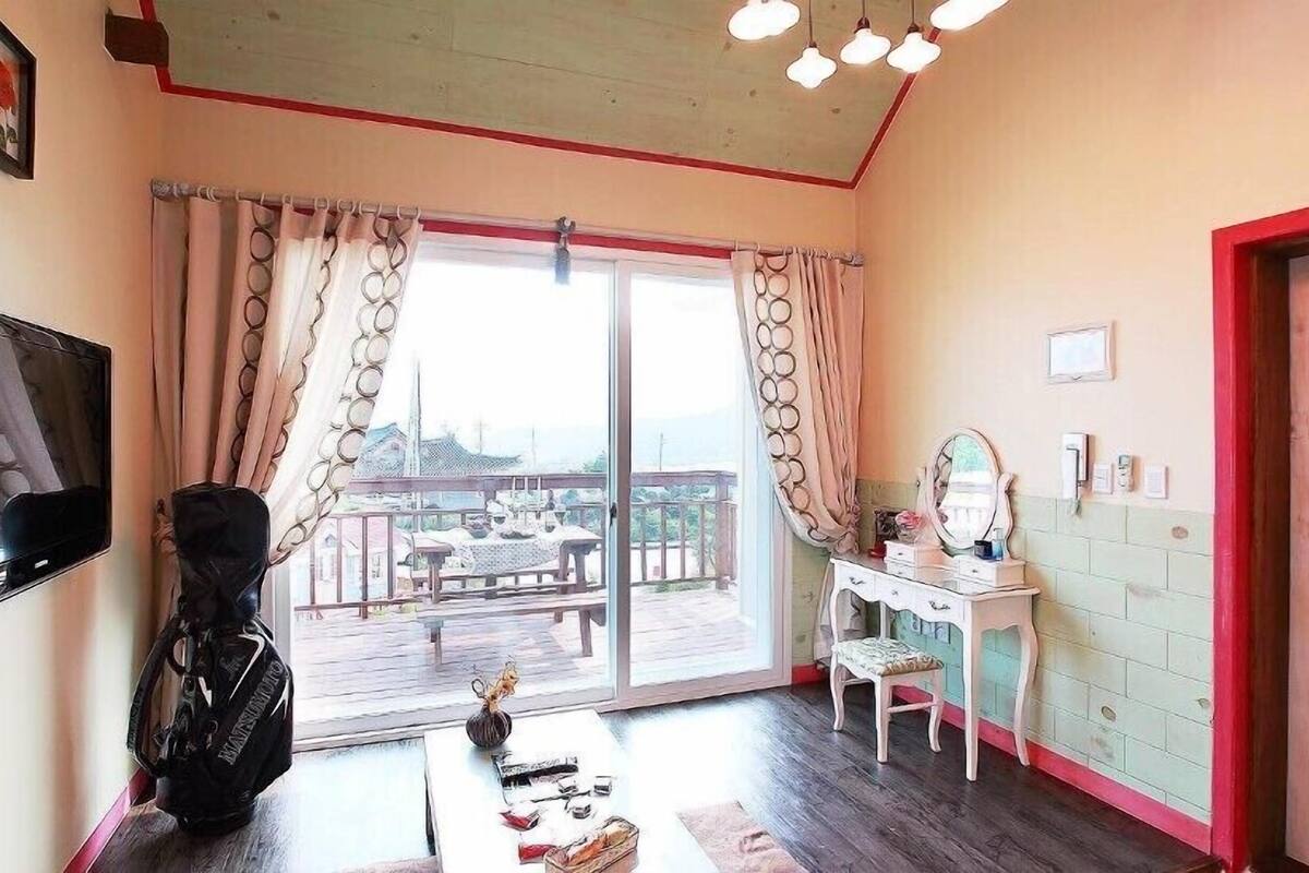 A room with a sliding glass door leading to a balcony, a desk with a chair, a mirror, and a wall-mounted TV.