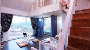 A spacious room with a large window, a bed, a table with a laptop, and a staircase.