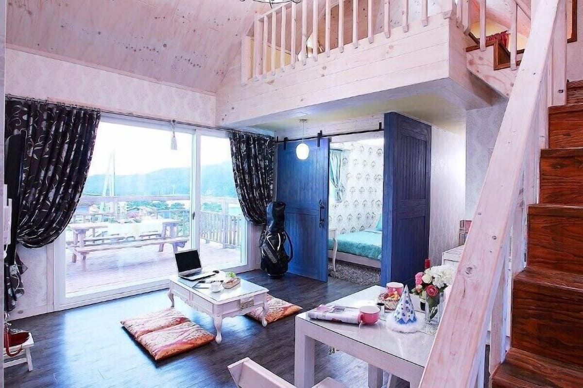 A spacious room with a large window, a bed, a table with a laptop, and a staircase.