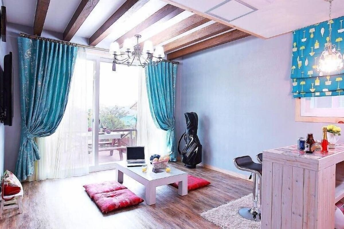A modern living room with a wooden ceiling, a TV mounted on the wall, a dining table with chairs, a red ottoman, and a small white cabinet.