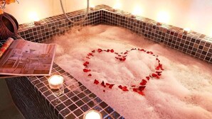 A bathtub with a heart-shaped arrangement of rose petals in the foam, surrounded by a tiled wall and a small candle.