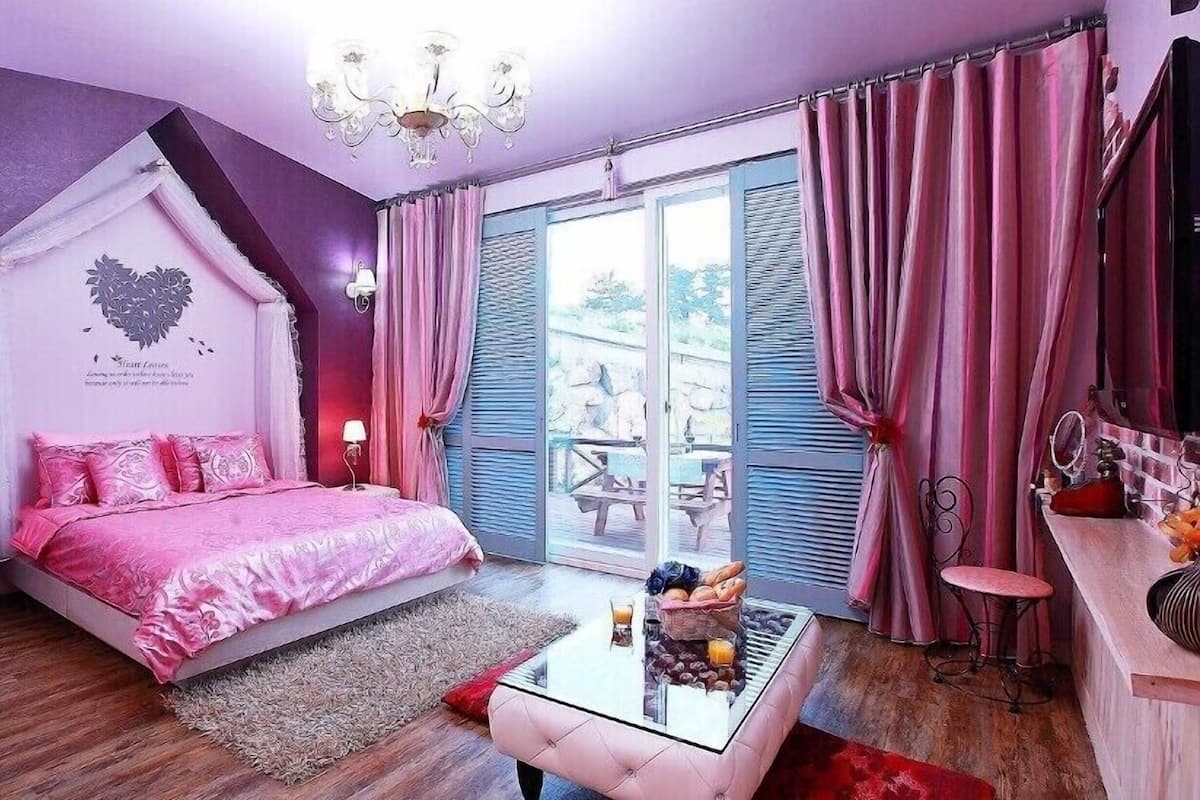 A bedroom with a bed, a glass coffee table, a chandelier, and a view of the outdoors.
