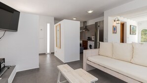 A modern living room with a sofa, a wooden bench, a flat-screen TV, and a kitchen in the background. - Holiday Home "Ca Sa Mola" with Terrace and Pool (Cala Blanca - Ciutadella de Menorca)