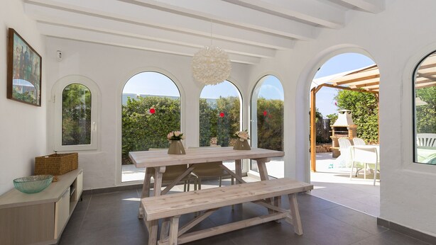 A dining area with a wooden table and benches, large arched windows, and a view of the outdoors. - Holiday Home "Ca Sa Mola" with Terrace and Pool (Cala Blanca - Ciutadella de Menorca)
