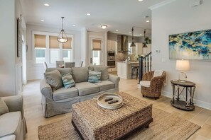 Un salon moderne avec un canapé, un pouf et des fauteuils, une cuisine en arrière-plan et un tableau au mur.