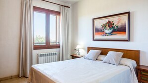 A bedroom with a large bed, a window with curtains, a radiator, a bedside table with a lamp, and a painting on the wall. - Rural Villa with pool and spectacular views – Villa Sastre (Campanet)