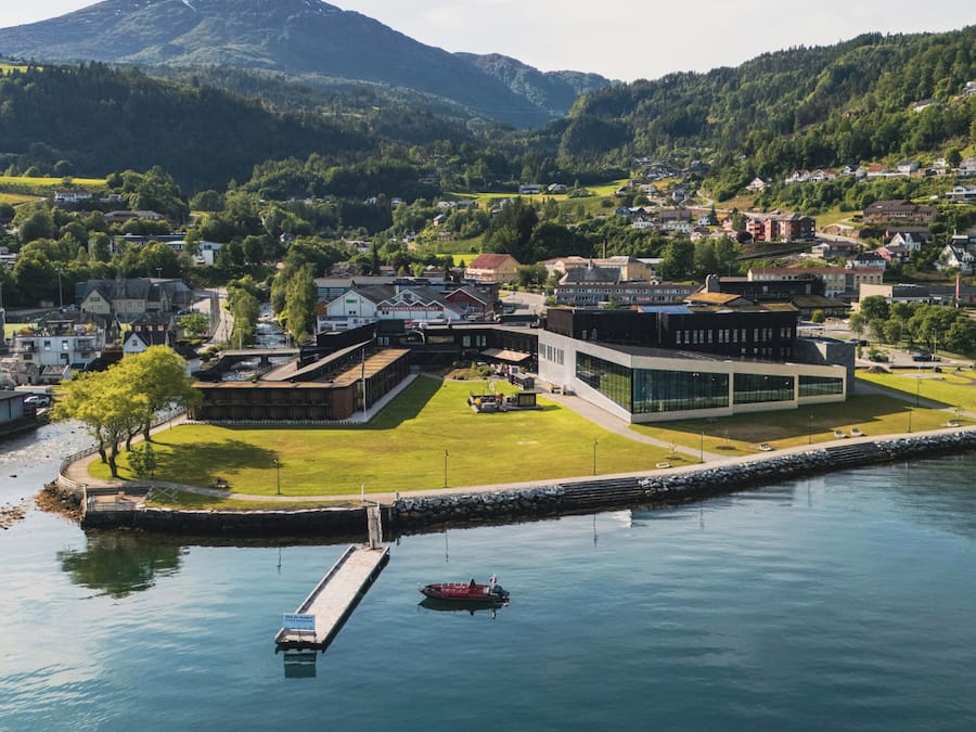 A modern building with a glass facade, situated by a calm body of water, surrounded by lush greenery and a mountainous backdrop.