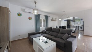 Uma sala moderna com um sofá seccional, uma mesa de centro de vidro e uma cozinha ao fundo.
